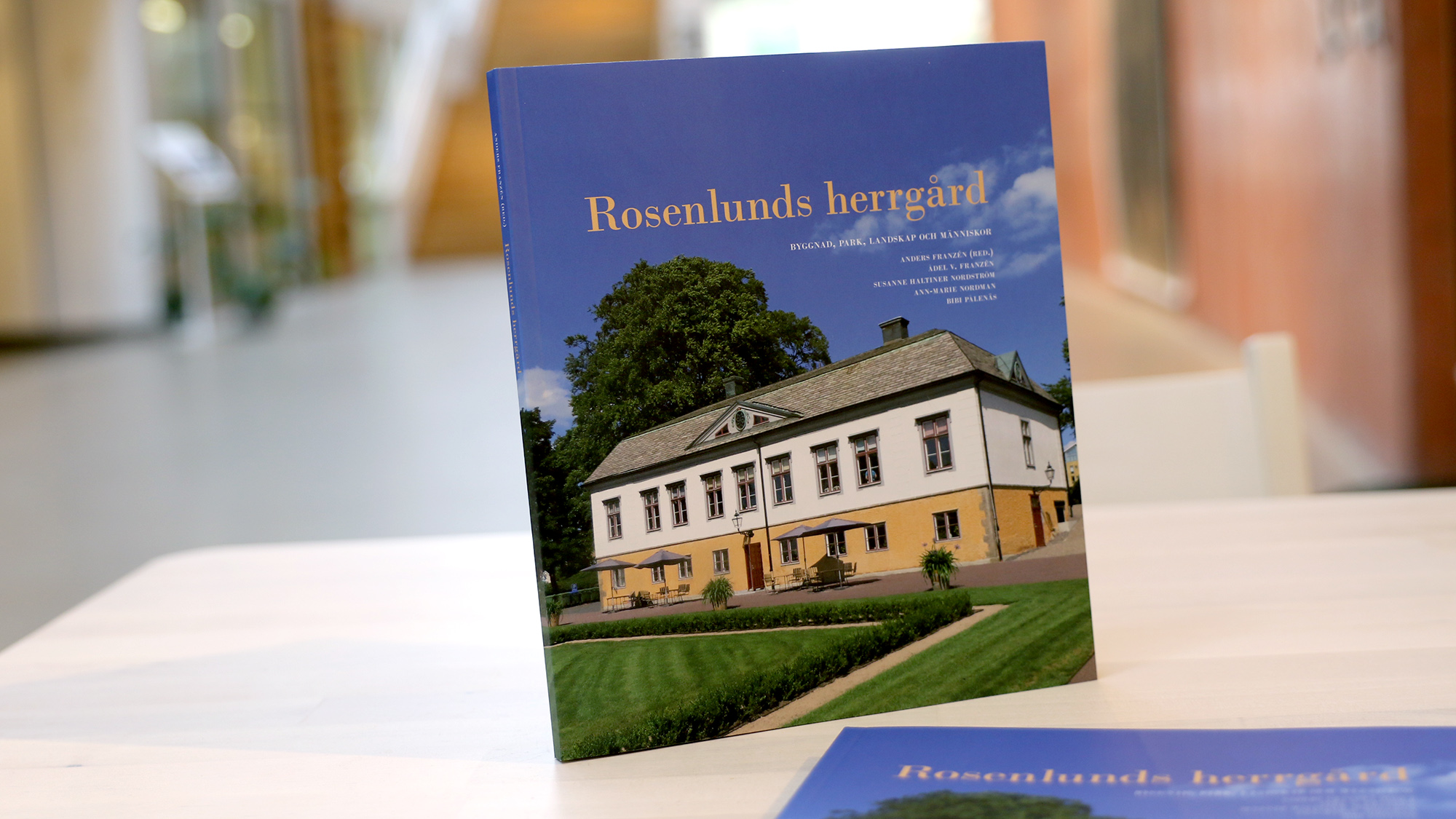 En bok står uppställd på ett bok inne i museets entréhall. Det står Rosenlunds herrgård på den i gult. Fotot på omslaget visar herrgårdsbyggnaden i sin helhet, med blå himmel bakom.