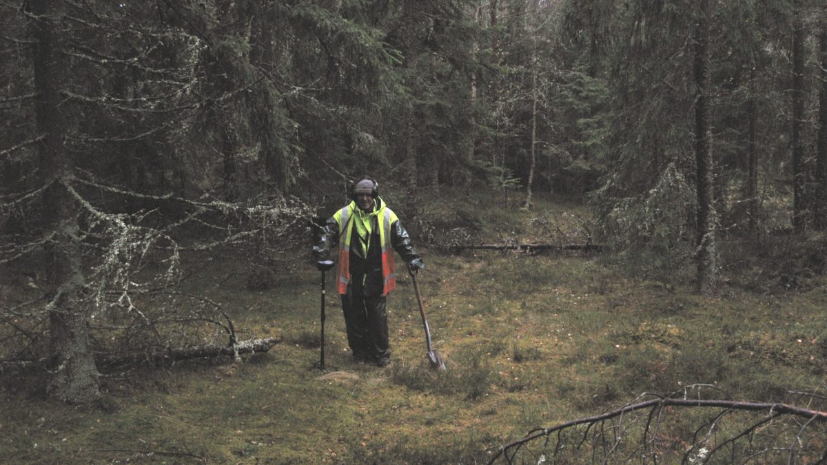 En arkeolog står med utrustning i regnet mitt i skogen.