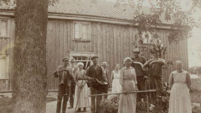 Svartvit gammal bild med kvinnor och män framför trähus.