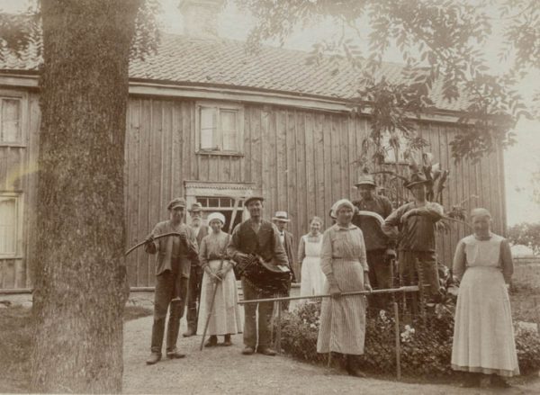 Svartvit gammal bild med kvinnor och män framför trähus.
