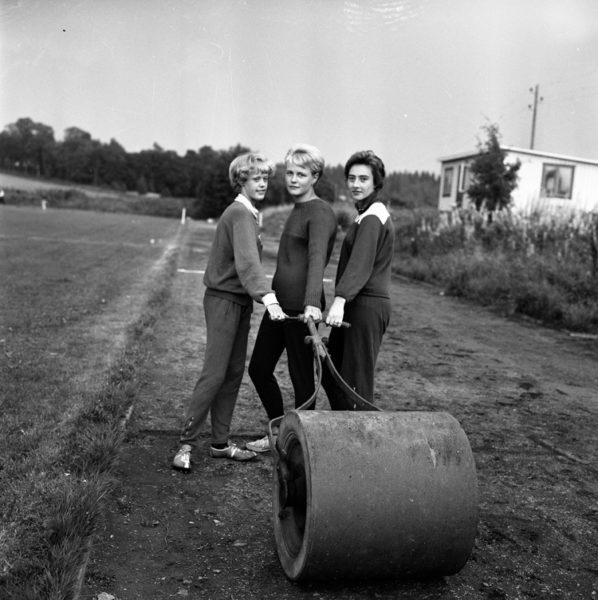 Svartvitt foto föreställande tre unga kvinnor som drar en vält på ett fält. Tidigt 1960-tal.