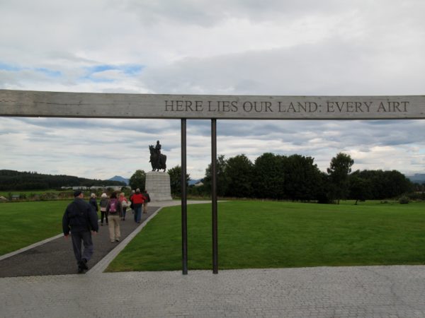 Monument med en person som sitter på en häst. Flera turister är påväg och ska kolla på monumentet. På en träbalk står orden "Here lies our land: every airt".