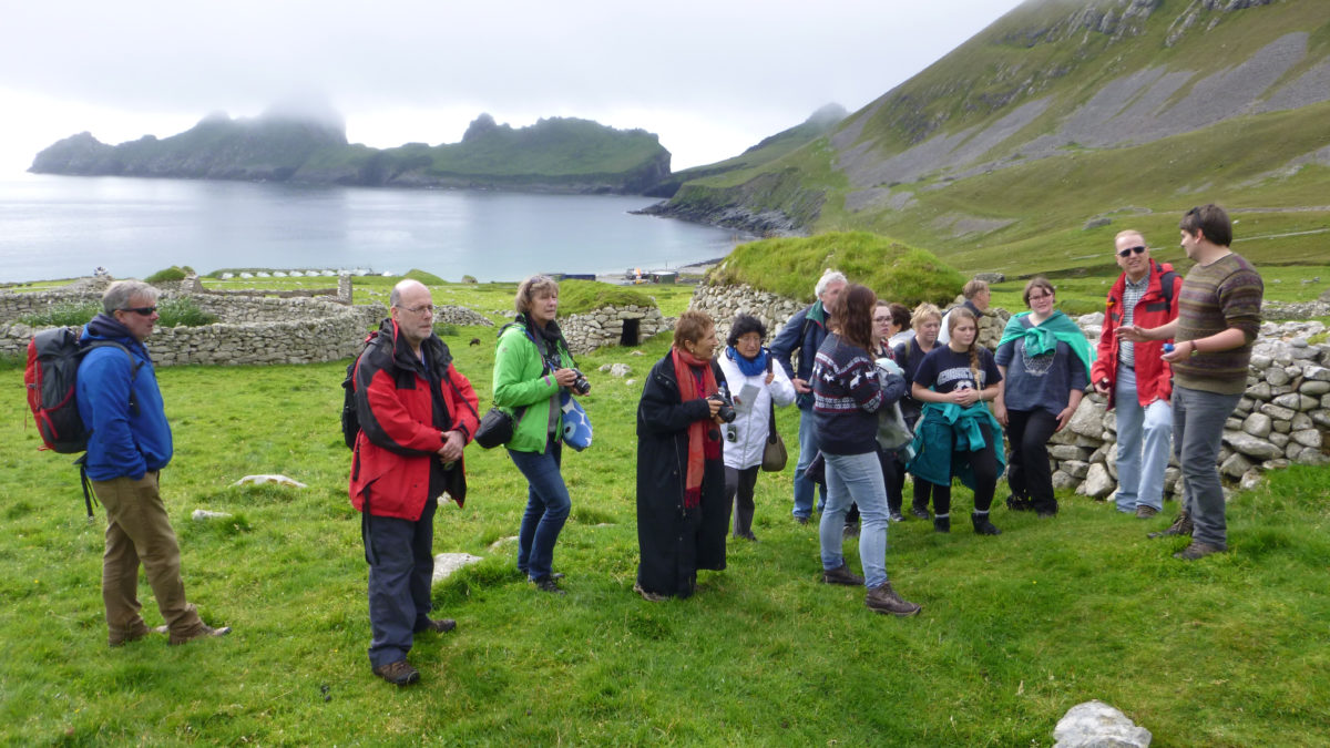En grupp med personer står och lyssnar på en arkeolog. Fin bakgrund med grönt gräs, hav, lite dis och bergiga delar.