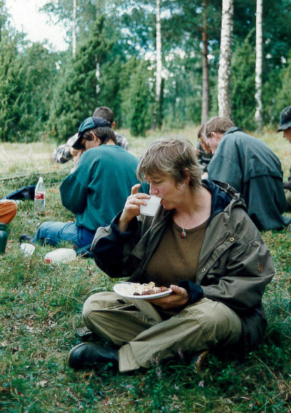 En kvinna främst, sitter och äter mat ute i naturen. Bakom sitter flera andra personer och gör detsamma.