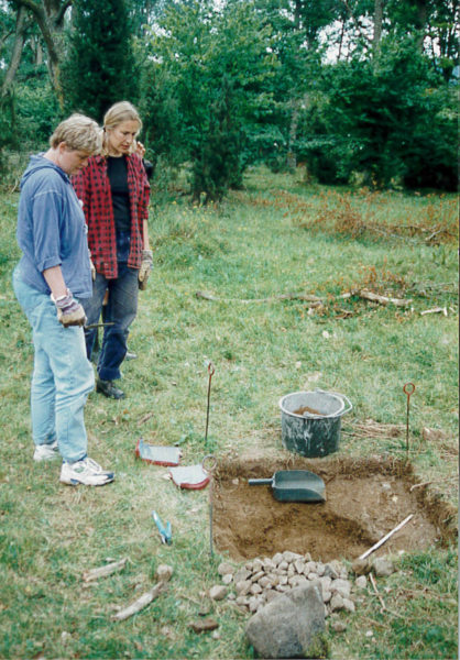 Två personer står vid ett uppgrävt hål. Redskap ligger runtom hålet.