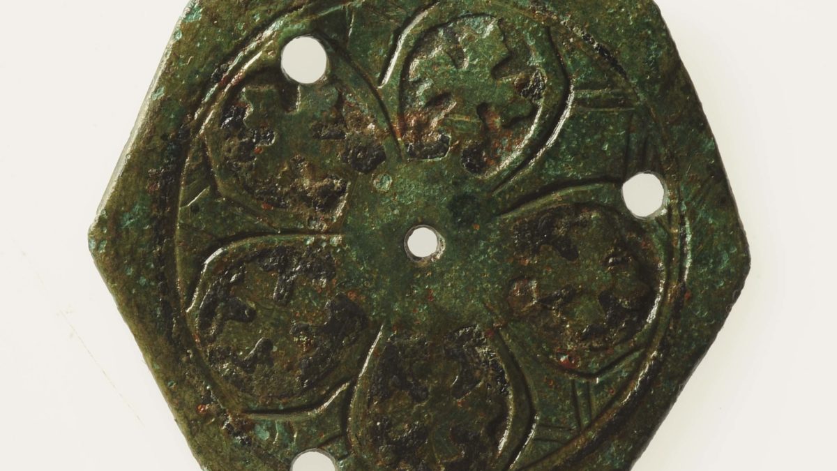 Ett åttakantigt bronsmärke med tre hål runt kanterna och ett hål i mitten. Märket har bladliknande inristningar.