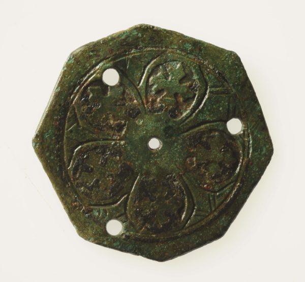 Ett åttakantigt bronsmärke med tre hål runt kanterna och ett hål i mitten. Märket har bladliknande inristningar.