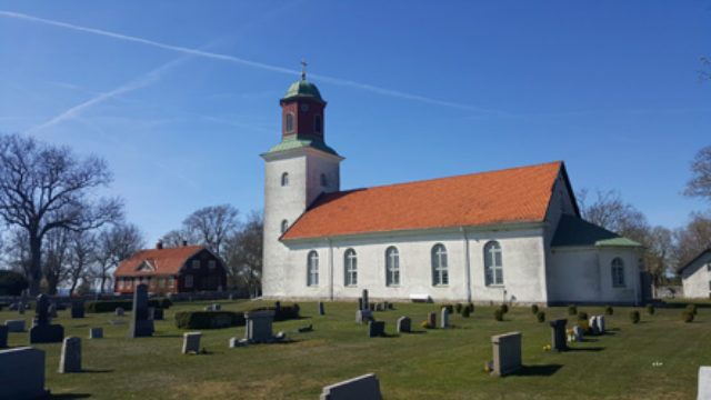 En ljus kyrka med tegelrött tak, gravstenar på kyrkogårdens gräsmatta framför.
