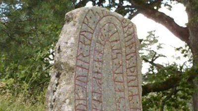 En stor sten med röd inristad skrift i sig. Stenen står i skogen bland gräs och träd.
