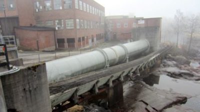 En fabrik med trasiga fönster. Bredvid fabriken ligger en flottningsränna och en stor tub av stål.
