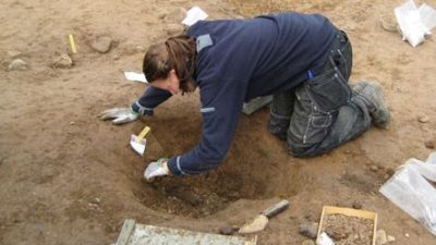 En kvinna sitter på knä och gräver i en grop. Runt omkring henne ligger en spade och plastfickor.