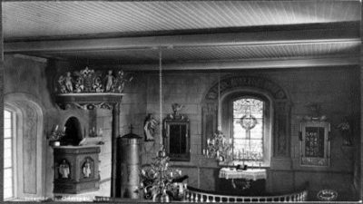 Gammal svartvit bild med föremål, rund järnkamin, altare och fönster.