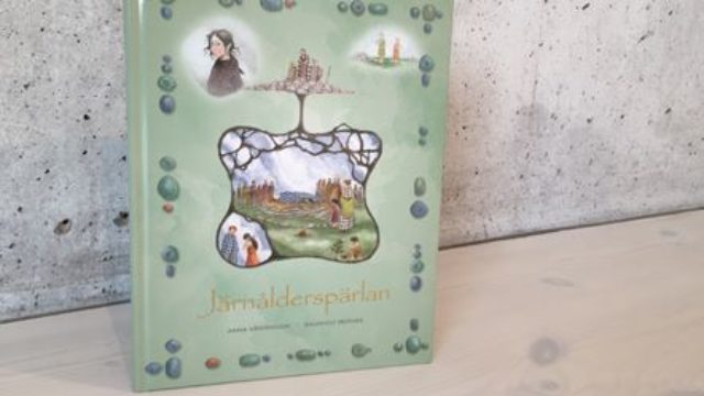 Bild av boken mot en grå vägg. Omslaget har titeln Järnålderspärlan mot en grön bakgrund. Akvareller föreställande barn i forntida klädsel. I mitten av bilden en illustration av en dåtida begravning. Allt är inramat av rader av pärlor.