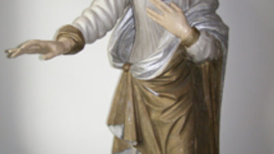 En stående kvinna med huvudduk och mantel. Hon håller en hand mot bröstet och den andra utsträckt.