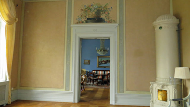 Vägg med dekorationsmålning. Till höger i rummet står en vit kakelugn.