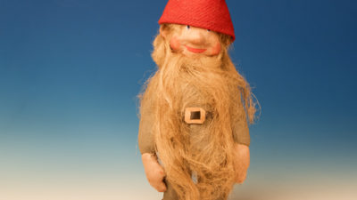 Jultomte-docka med skägg, gråa kläder och röd luva, skärp och skor av skinn.