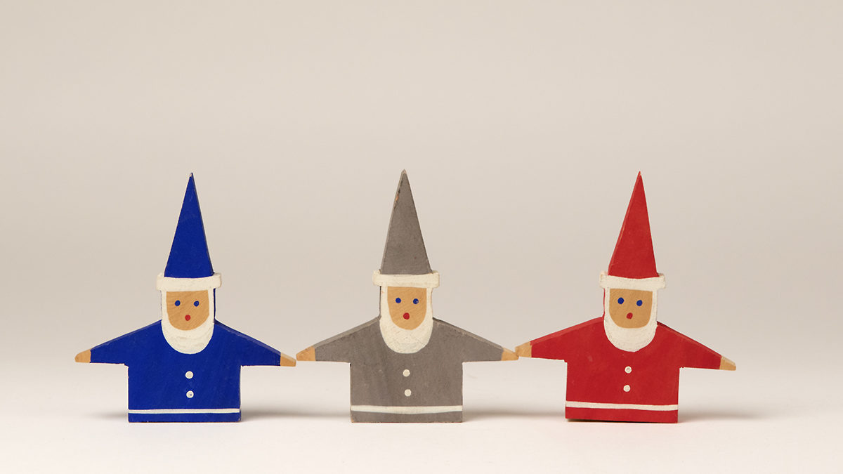 Tre jultomtar stående bredvid varandra, målade i respektive färg blått, grått och rött. Formen är en platt figurskulptur av trä med endast överkropp. Armarna är utsträckta åt sidorna. På huvudet sitter en upprättstående spetsig mössa.