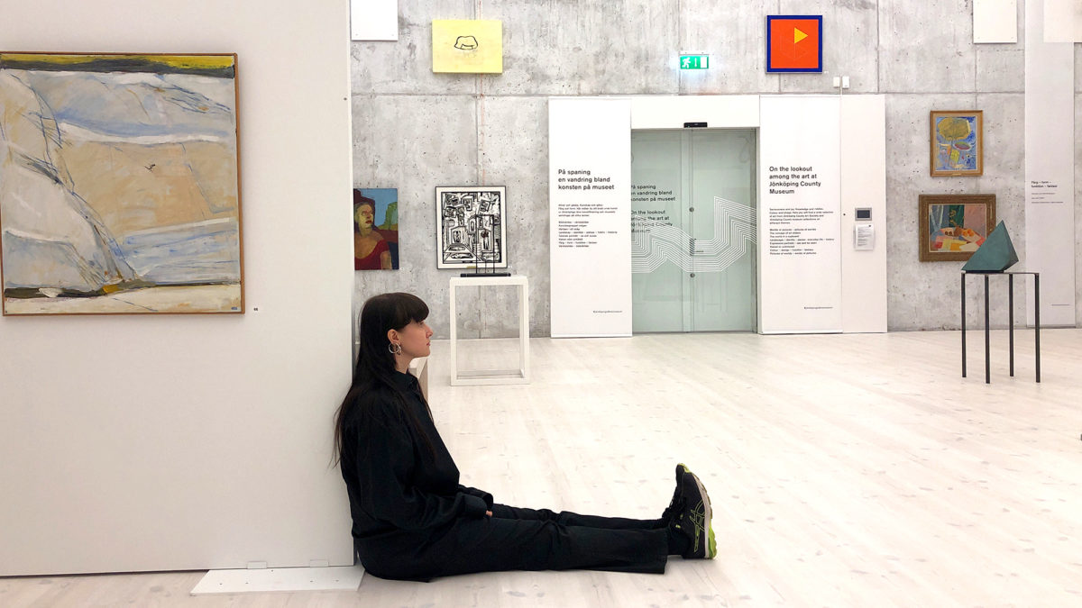 En kvinna i svarta kläder sitter på golvet i en stor ljus utställningssal. Hon lutar sig mot kanten av en vägg. Man anar tavlor och föremål omkring henne.
