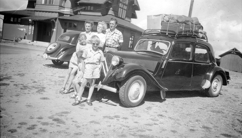 Bilsemester. Tre vuxna personer och tre barn står framför en bil med packning fastsurrad på taket.