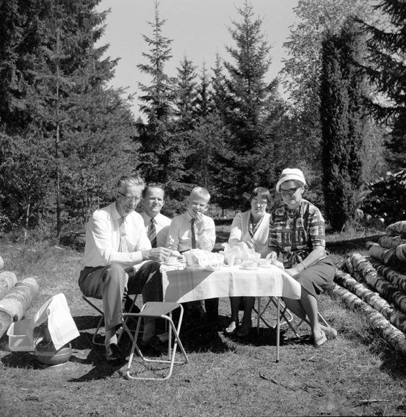 Fem personer sitter runt ett dukat bord intill en skog.