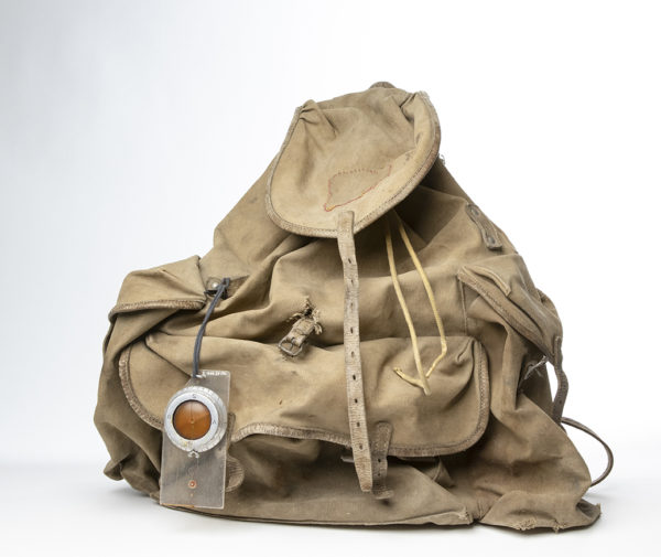 Äldre modell av ryggsäck med päronformad säck av kraftigt grå-brunt tyg. På ryggsäcken hänger ett kompass.