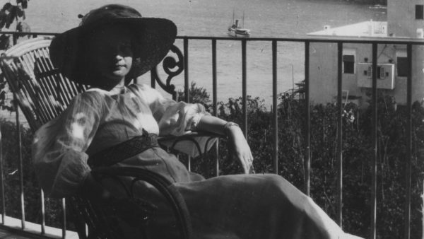 Svartvitt foto av Ester Bauer med solhatt, sittandes på en balkong/verande med utsikt över havet