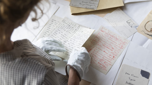 Äldre brev utspridda på ett bord. Person med handskar håller i ett av breven.