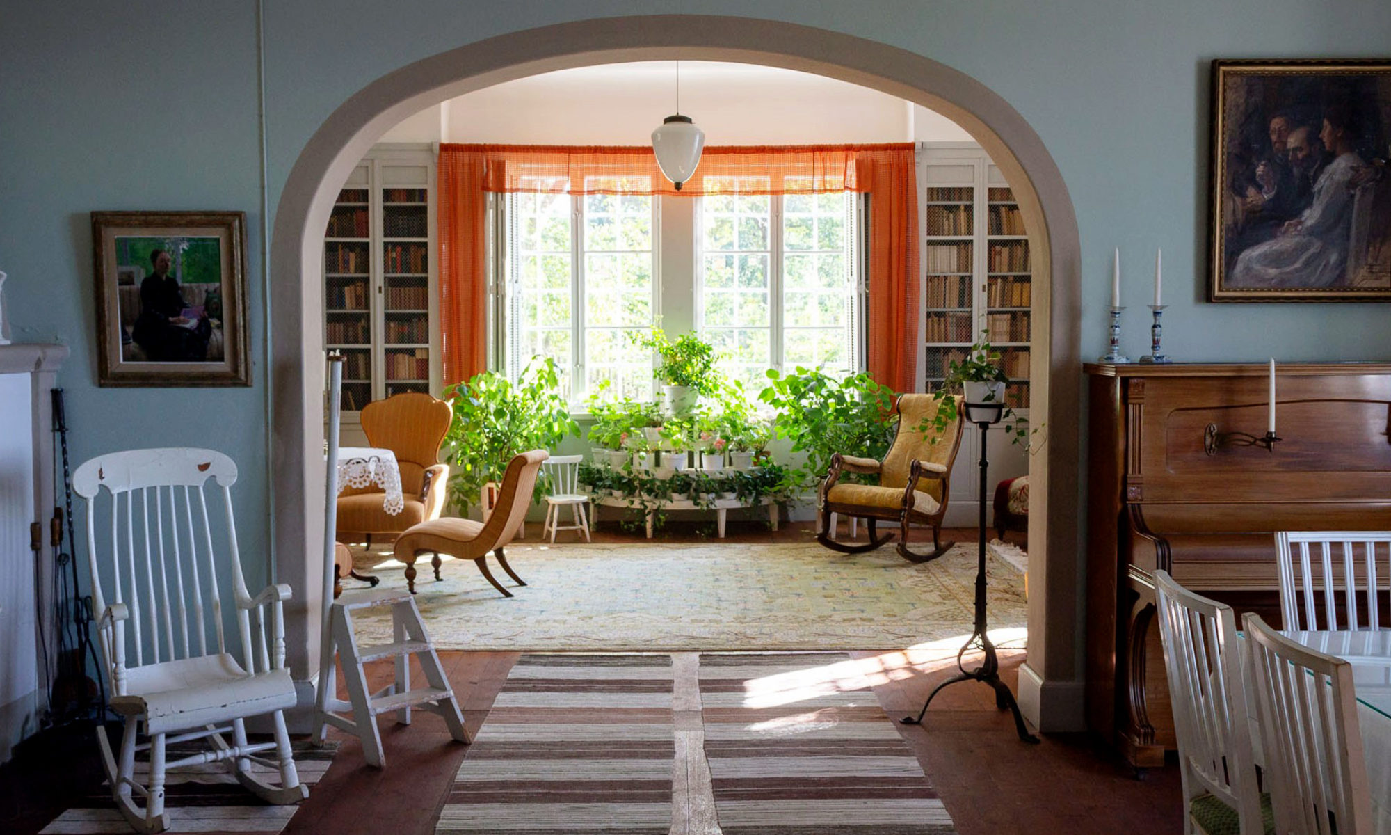 Ett stort rum med ett valv syns rakt fram, roströda gardiner och grönskande krukväxter i fönstret rakt fram. Trasmattor på golvet.