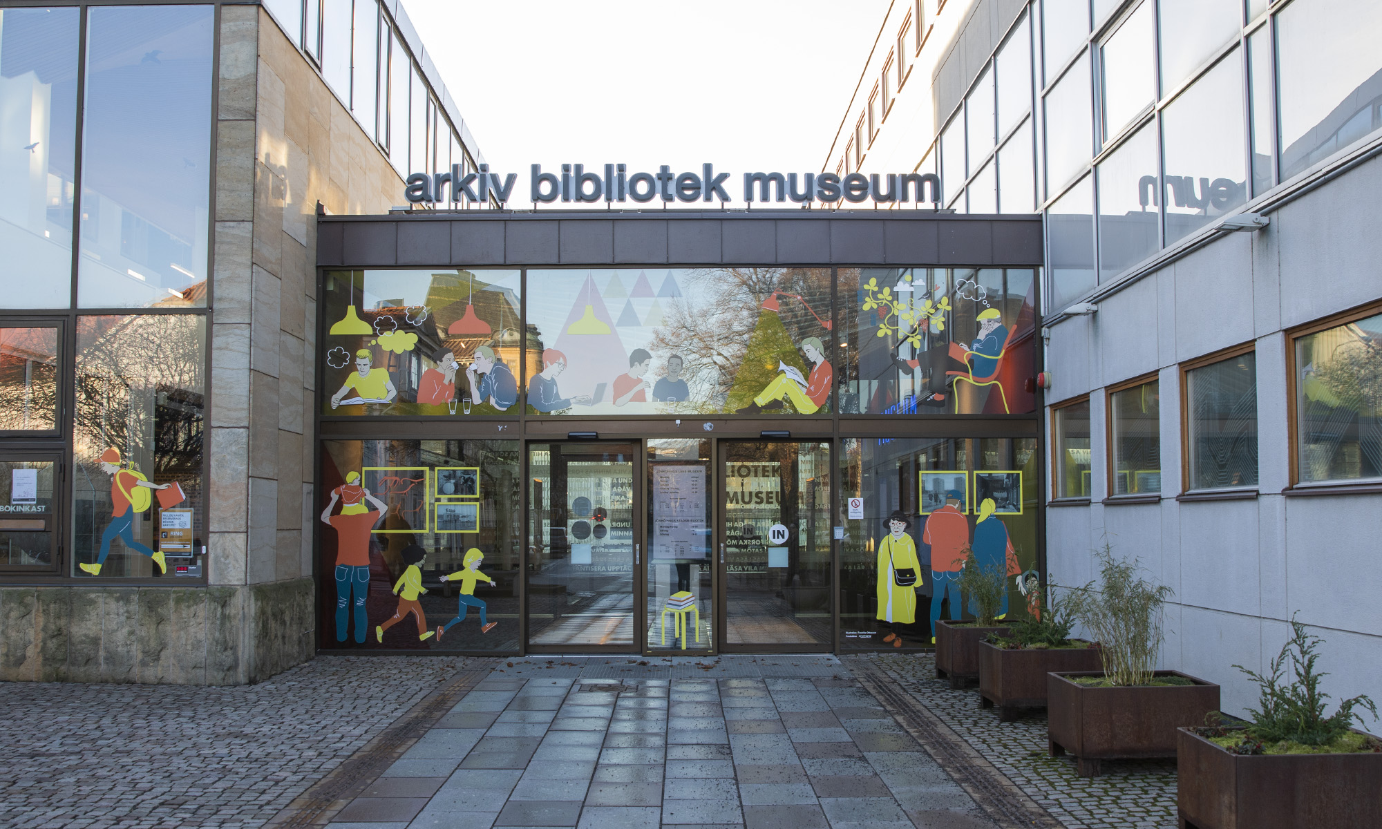 Grå byggnad med glaspartier på framsidan. Ovanpå taket sitter bokstäver som bilder orden arkiv, bibliotek, museum,.