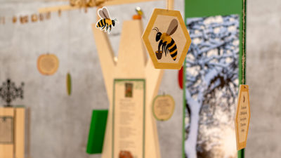 En utställning där träd med utställningstexter och föremål står. I luften hänger små skyltar i form av bin och äpplen.