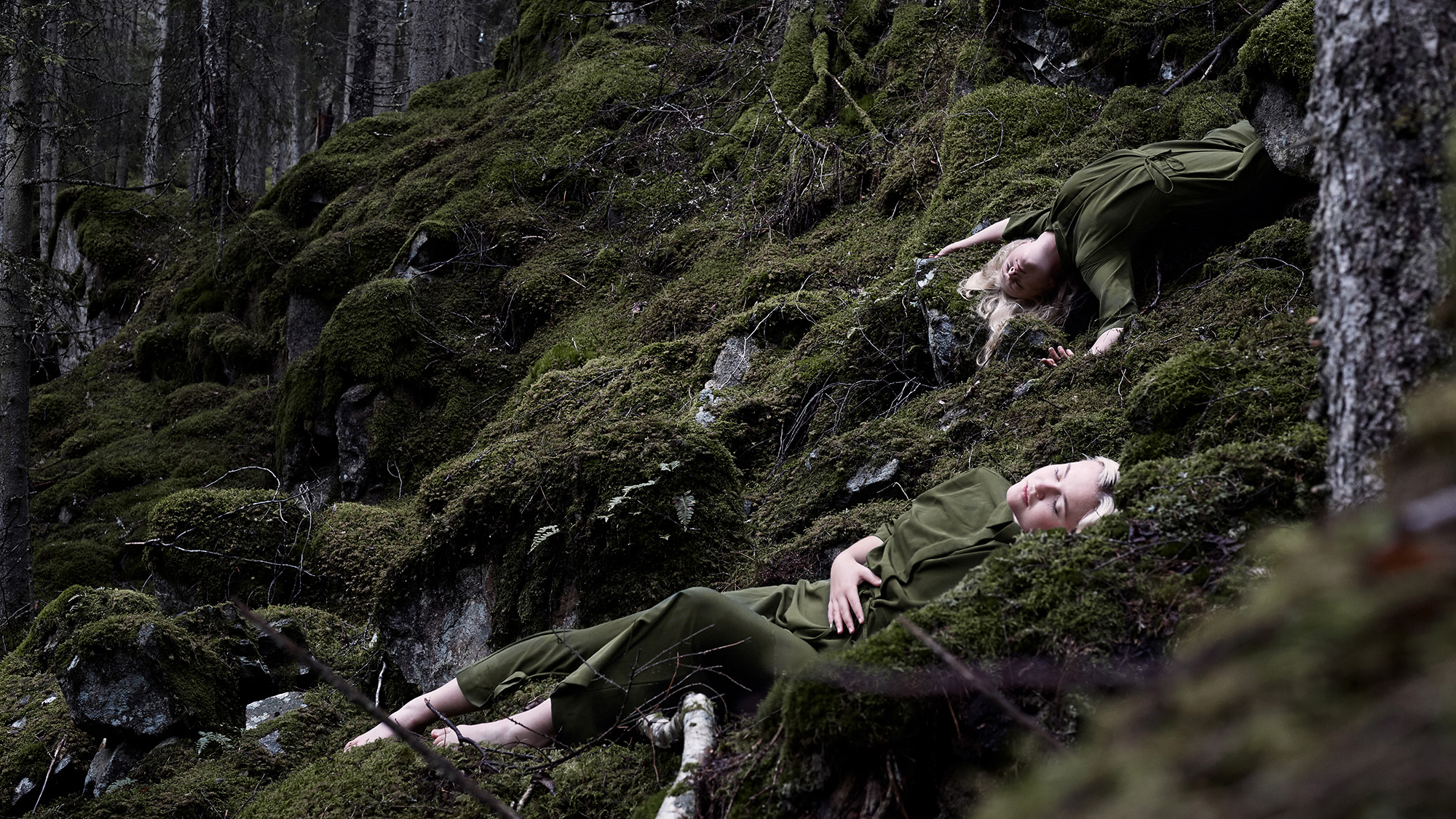 Två unga kvinnor ligger på mörkgrön mossa i en skog. De blundar och är liksom omfamnade av den omgivande naturen.
