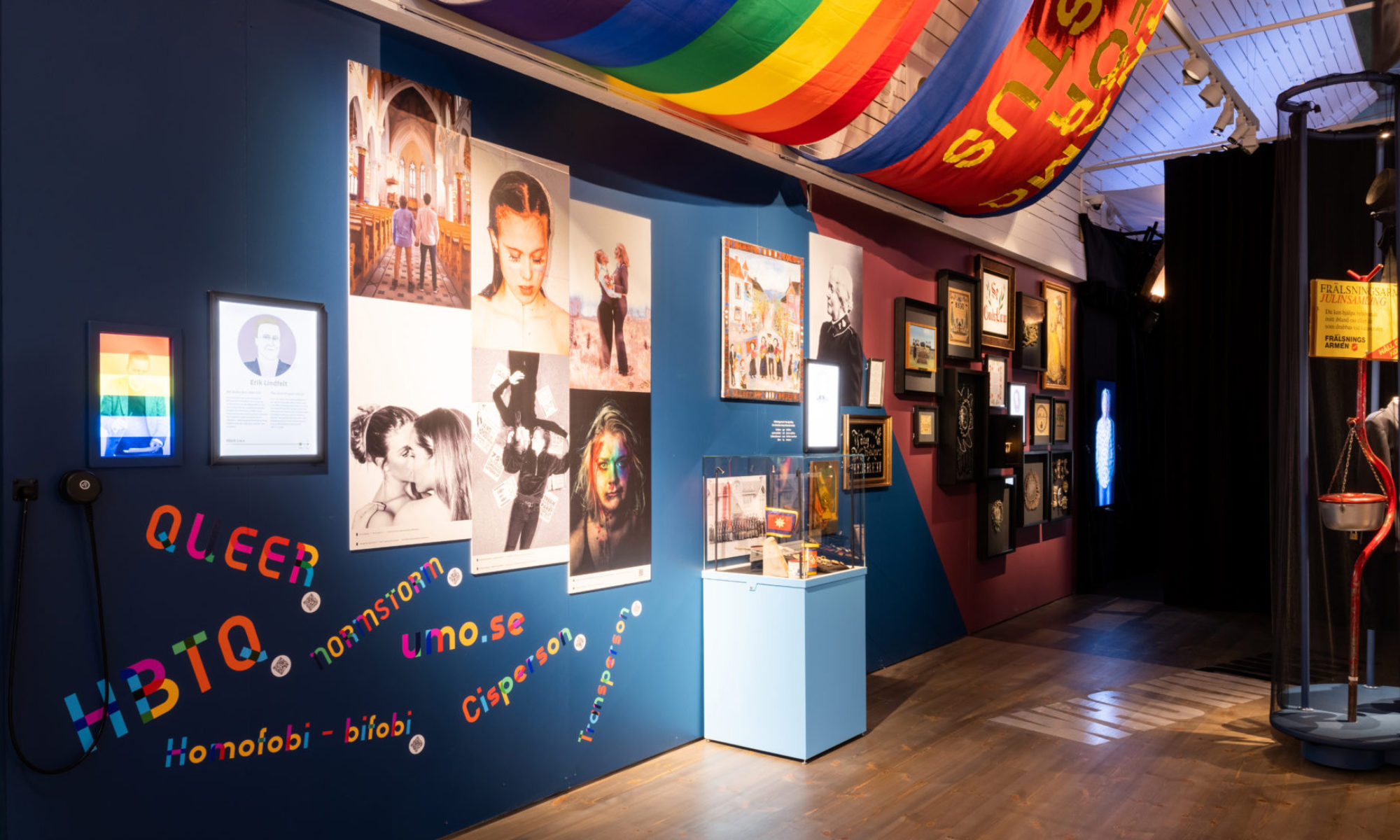 Foton och föremål syns på en blåmålad vägg, där också ord som rör HBTQ-frågor är uppsatta. I taket syns bland annat en regnbågsflagga.