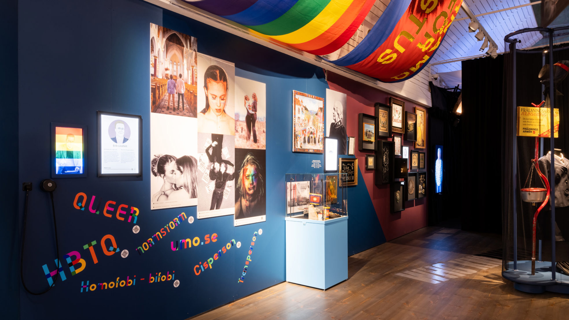 Foton och föremål syns på en blåmålad vägg, där också ord som rör HBTQ-frågor är uppsatta. I taket syns bland annat en regnbågsflagga.