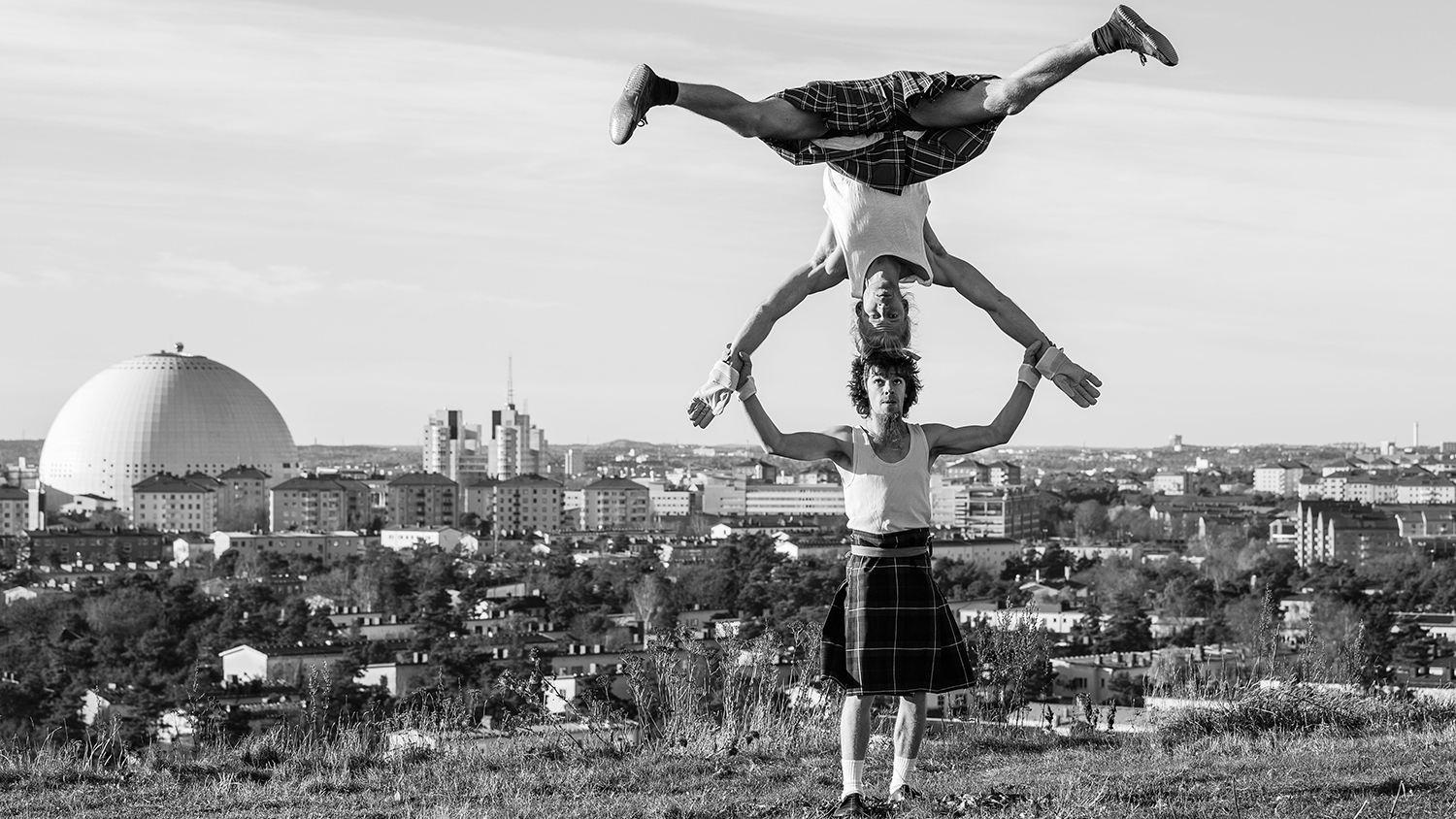 Två cirkuskonstnärer balanserar. Den ena personen står på huvud mot den andras huvud, de håller varandra i händerna. I bakgrunden syns utsikten över Stockholm med Globen till vänster i bild.
