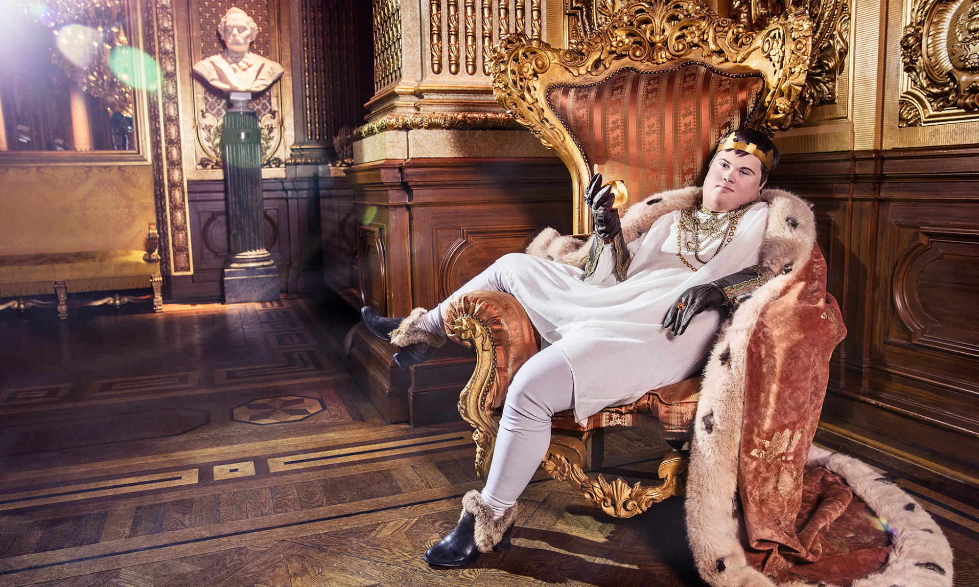 En person föreställande en klassisk kung sitter avslappnat i en tron med ena benet på armstödet. Bakgrunden innehåller detaljer av guld, rött och sammet.