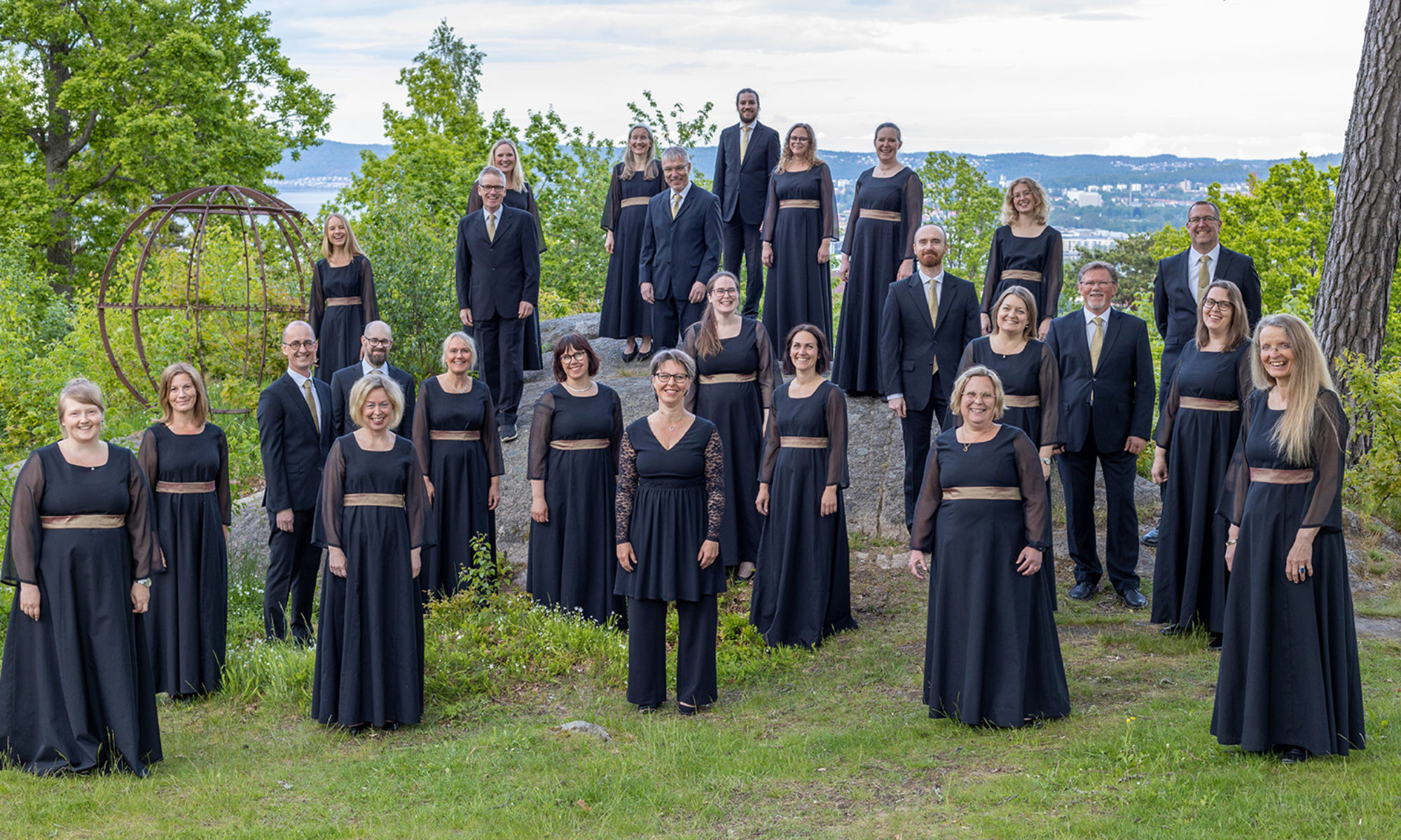 Ett 20-tal personer klädda i svarta enhetliga kläder står uppställda på olika nivåer omgivna av grönska mot en utsikt över Jönköping