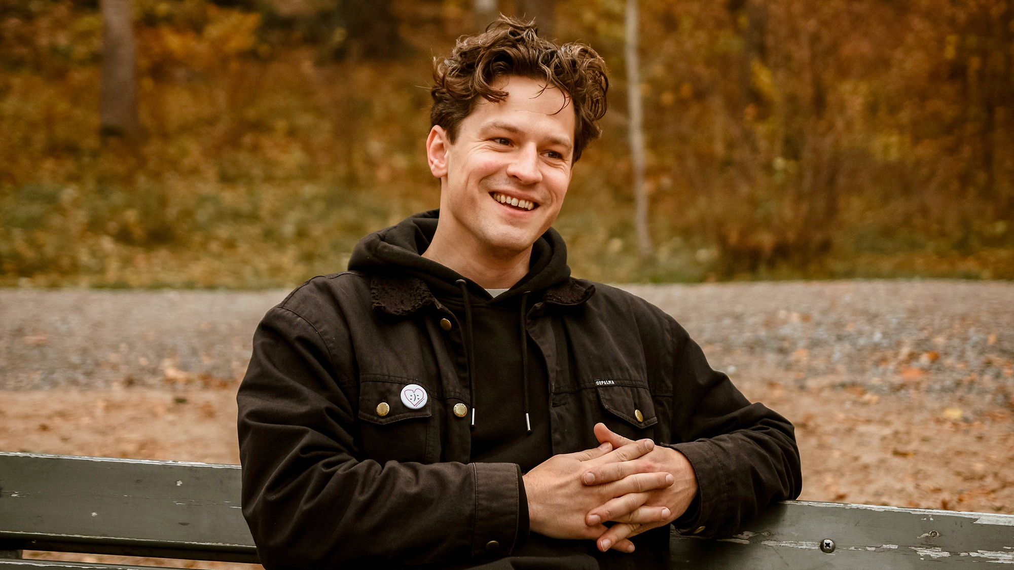 Porträtt av Henrik Wahlström. Han sitter utomhus på en träbänk, blickar åt sidan och ler. Iklädd svart jacka och 
