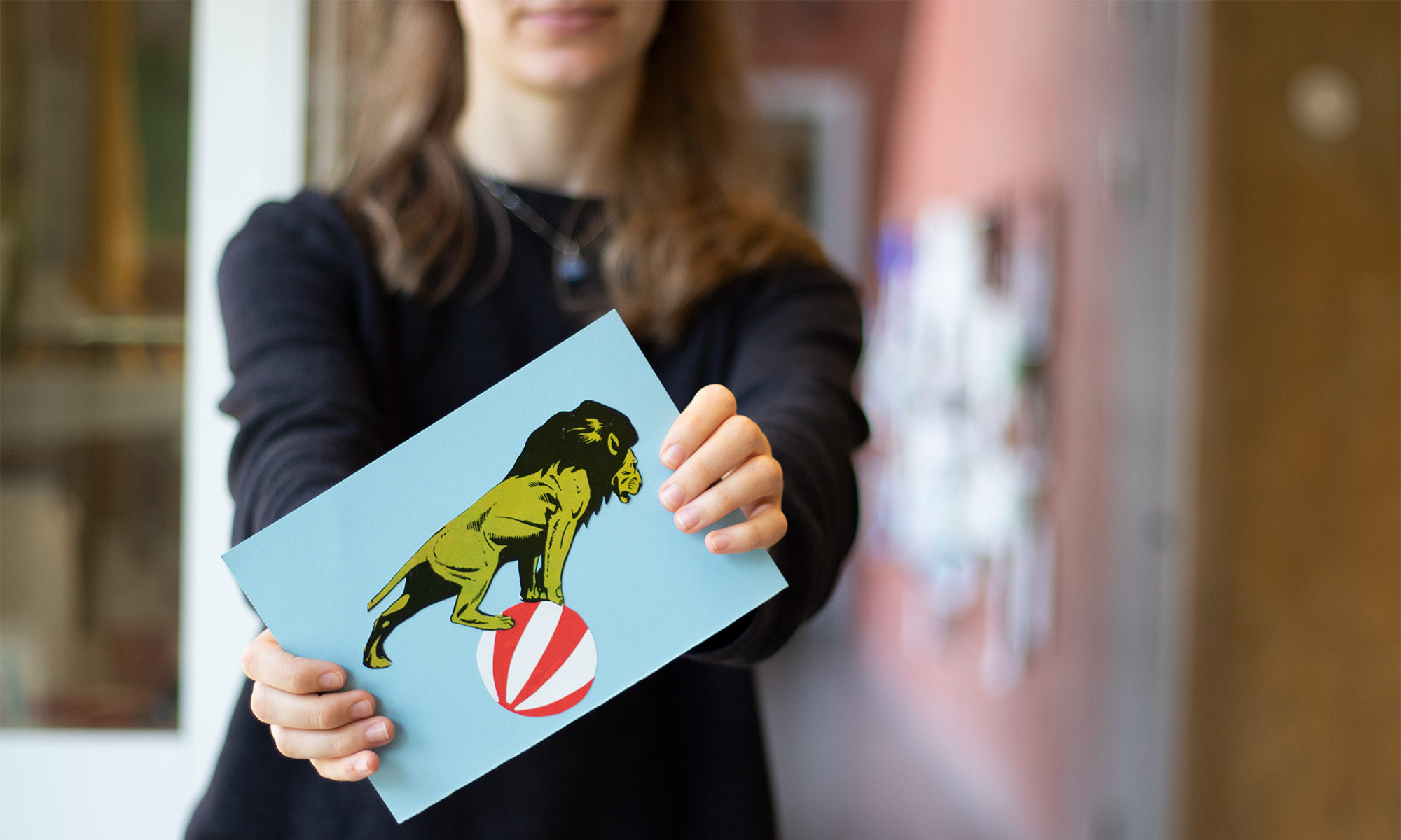 En person med långt hår och mörk tröja håller fram ett pappersark mot kameran. Pappret är ljusblått med bilden av ett grönt lejon stående på en rödvitrandig badboll.
