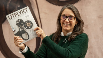 Redaktionssamordnare Patricia Monroy håller i ett nummer av tidskriften Utflykt med båda händerna. Hon ler mot kameran. I bakgrunden syns numrets omslag stort på en vepa på väggen. Det är kugghjul och drivhjul i mörkbrunt.