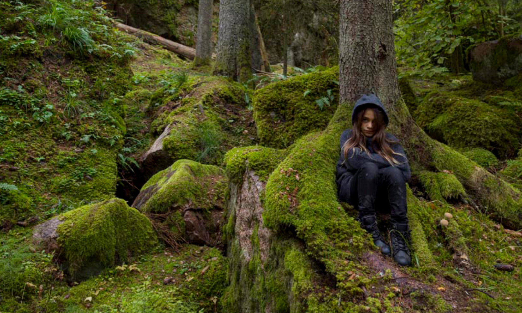 En ungdom sitter i en mossig skog omgiven av stenar och träd. Hen har en luvtröja med luvan uppdragen.