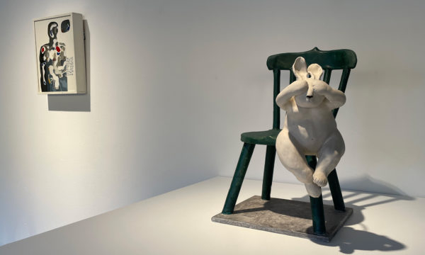 konstverk föreställande en vit kanin som håller för ögonen och hoppar från en grön stol. I bakgrunden anas ytterligare konstverk.