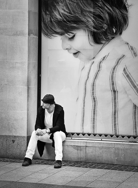Svartvitt foto av en sittande man och i bakgrunden en stor affisch av ett barn med blicken nedåt
