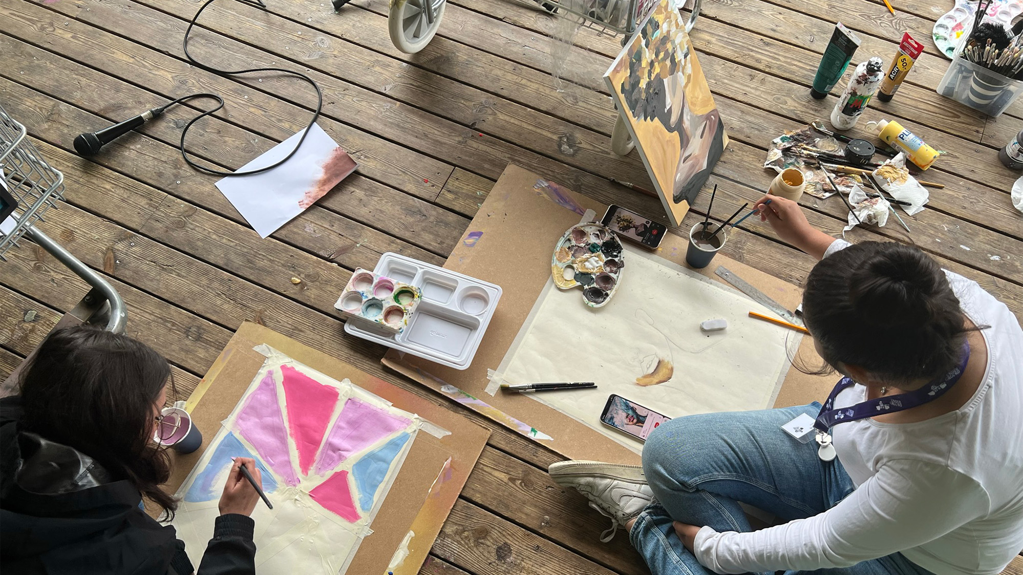 Två personer sitter på ett trägolv och målar med penslar och färgglada färger