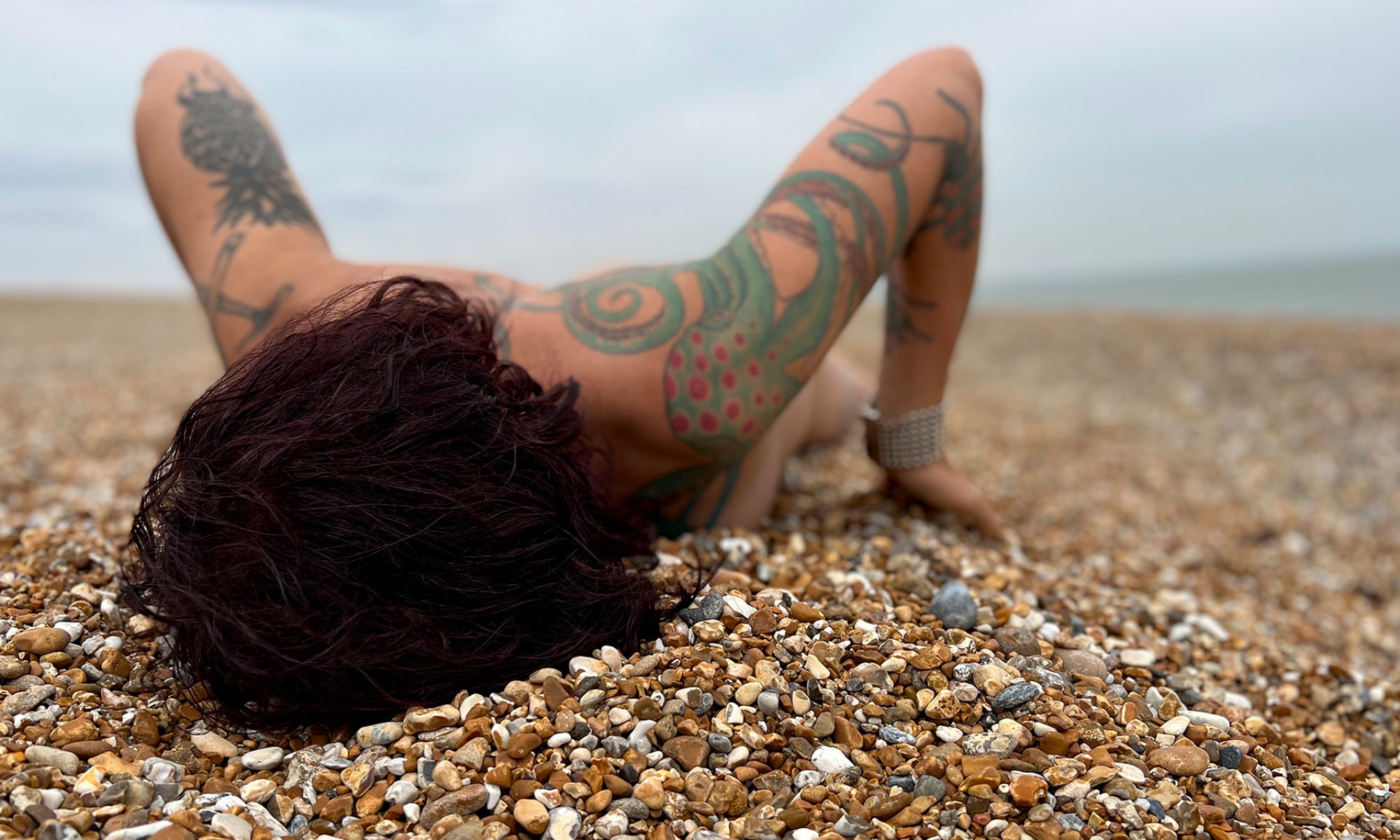 En person ligger på en strand täckt av småsten, med ansiktet nedåt. Armarna har tatueringar.