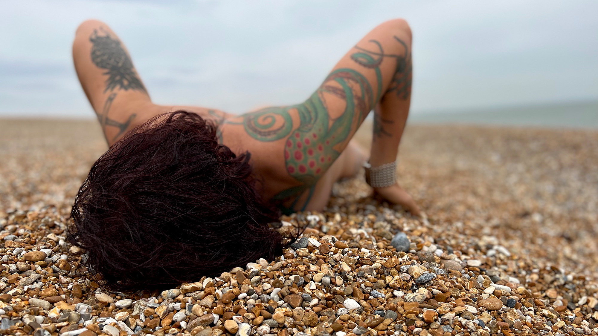 En person ligger på en strand täckt av småsten, med ansiktet nedåt. Armarna har tatueringar.