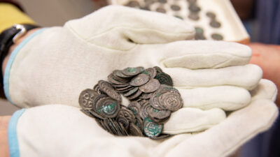 En samling mynt hålls av en person iklädd vita handskar. I bakgrunden en ask med fler mynt.