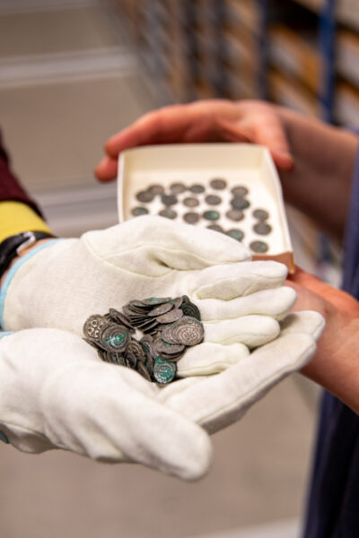 En samling mynt hålls av en person iklädd vita handskar. I bakgrunden en ask med fler mynt.