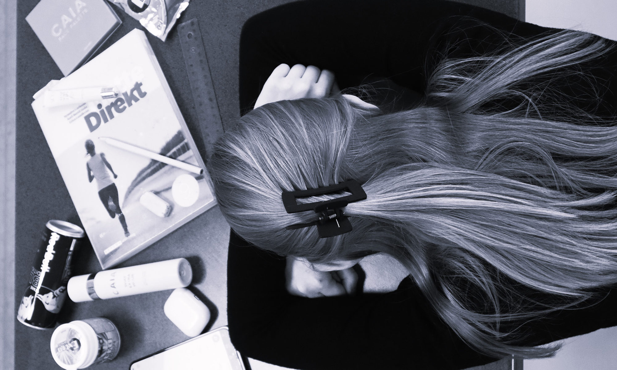 Person med långt hår ligger med sitt huvud mot sina händer på ett skrivbord fullt med böcker och saker.