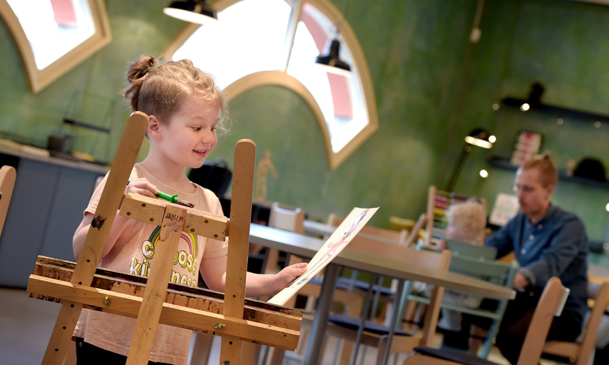 Ett barn vid ett staffli håller i en egengjord målning. I bakgrunden syns en vuxen och ett barn som läser