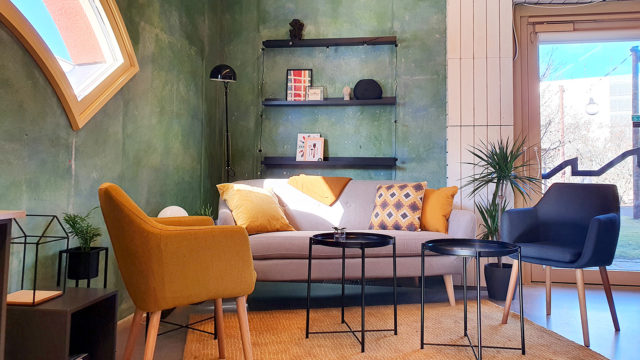 Varma färger på fåtölj, soffa, hyllor och brickbord mot en grönmålad betonvägg